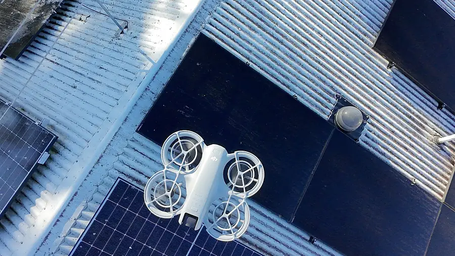 A small drone hovering over a metal roof with solar panels during a roof inspection